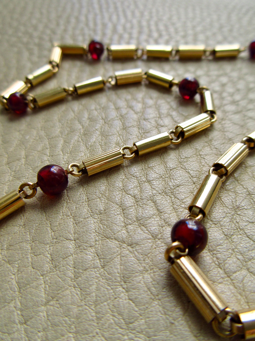 vintage 18k gold chain necklace featuring cylindrical links and dark red garnet beads, displayed on a textured beige leather surface.