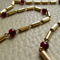 vintage 18k gold chain necklace featuring cylindrical links and dark red garnet beads, displayed on a textured beige leather surface.