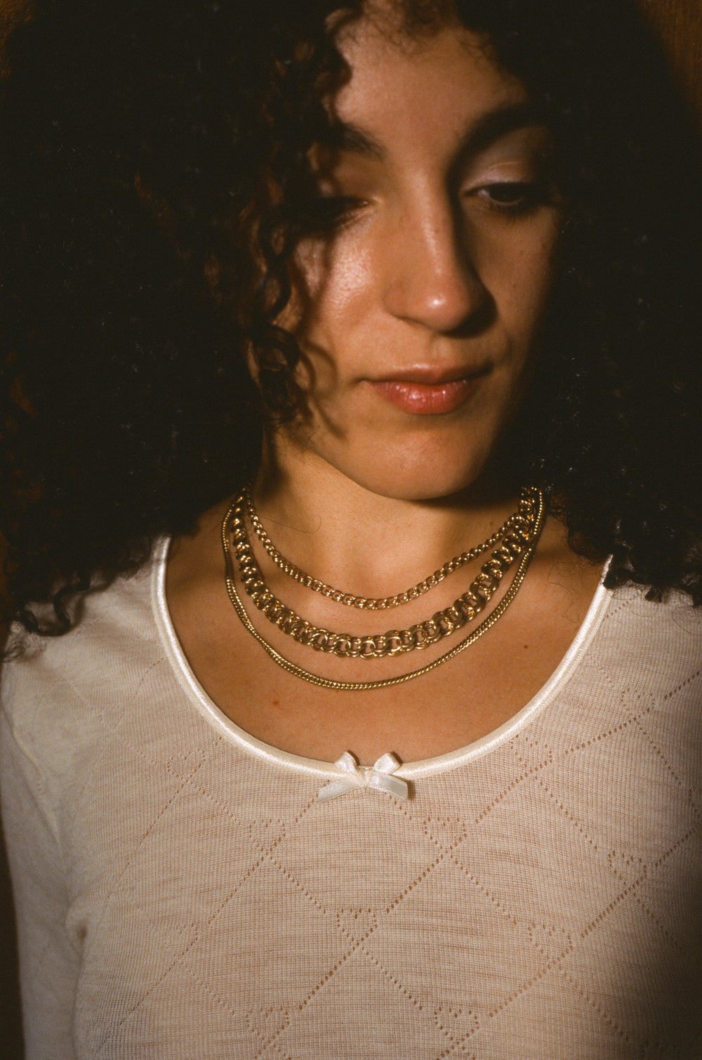 A close-up portrait of a woman with curly dark hair wearing layered gold necklaces over a cream-colored knit top.