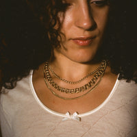 A close-up portrait of a woman with curly dark hair wearing layered gold necklaces over a cream-colored knit top.