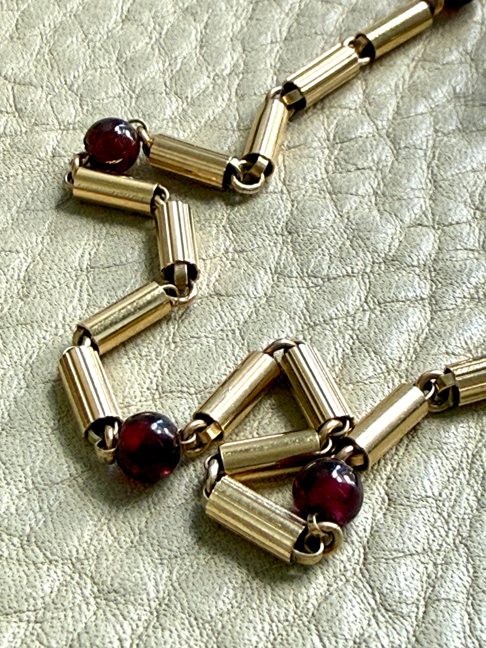 A close-up of an 18k gold chain necklace with cylindrical links and dark red round garnet gemstone accents, laid on a textured light beige surface.