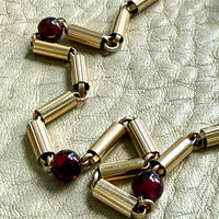 A close-up of an 18k gold chain necklace with cylindrical links and dark red round garnet gemstone accents, laid on a textured light beige surface.