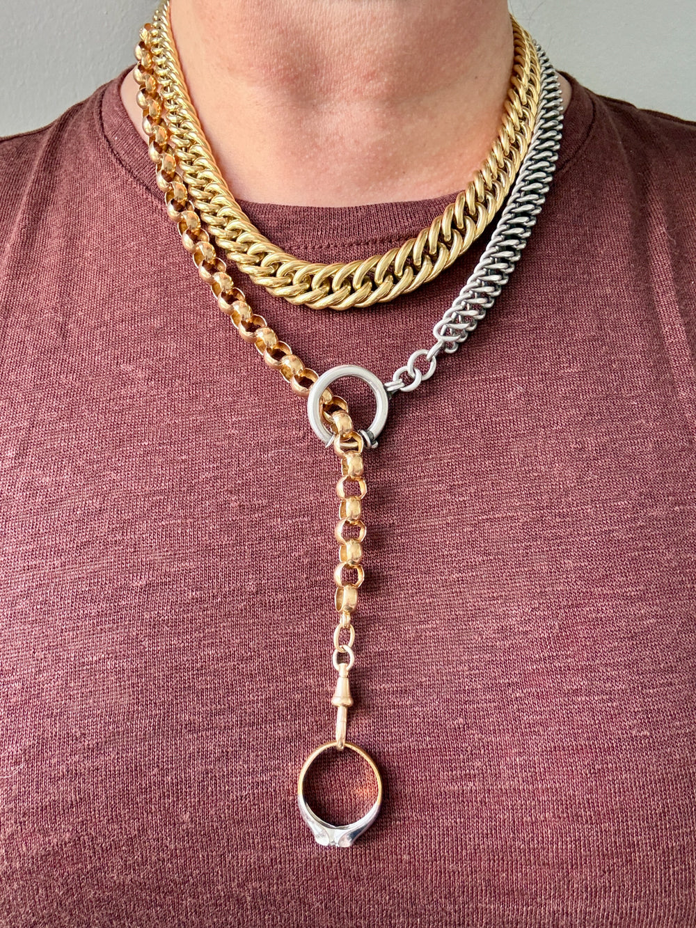 A close-up of a person wearing a layered necklace set featuring a thick gold Cuban link, a silver-toned chain, and a mixed gold and silver Y-necklace with a dangling ring charm.
