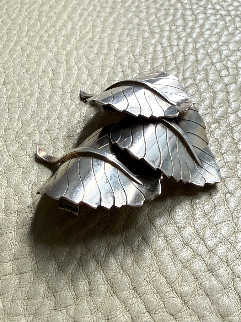 A sterling silver metal brooch shaped like overlapping leaves, photographed on a textured light beige leather surface.