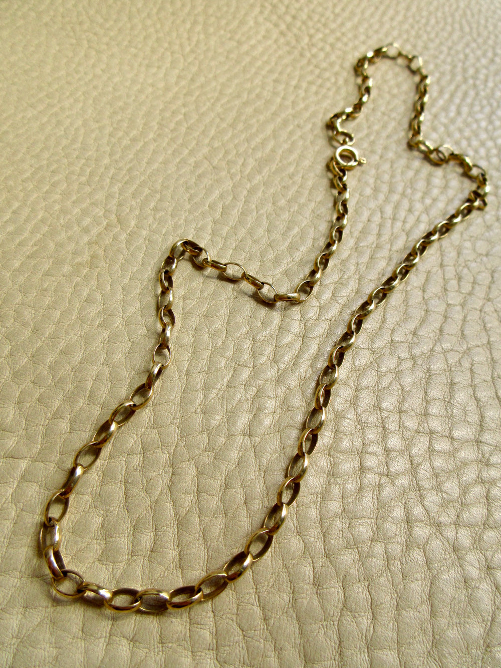 An 9k gold chain necklace with a twisted oval link design, laid out on a textured beige leather surface.