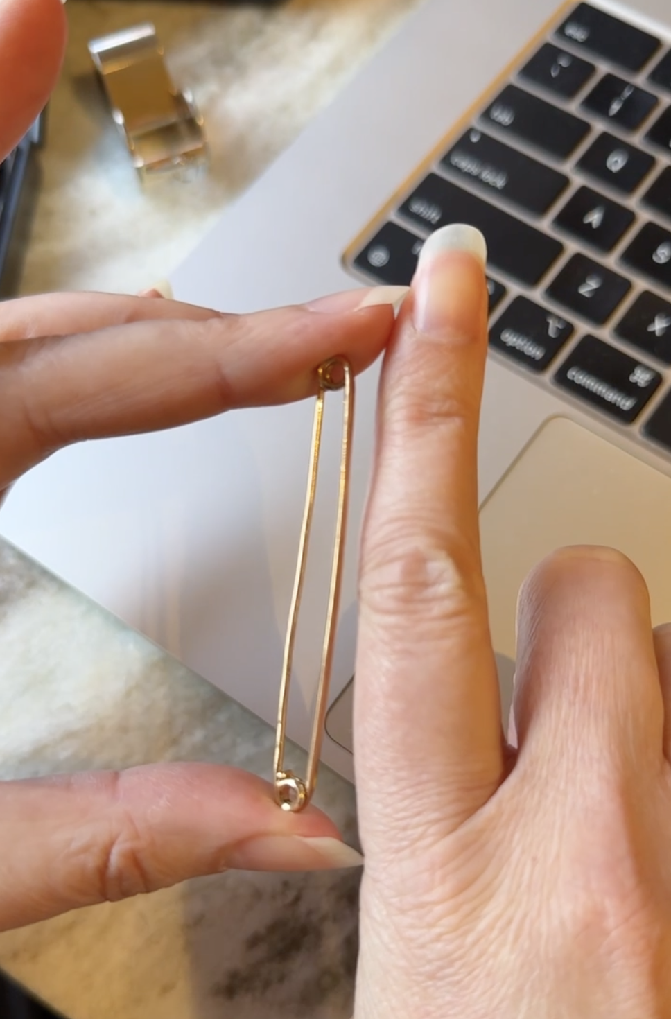 Gold safety pin held between fingers with a laptop in the background